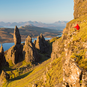 a person in a red jacket sitting on a rocky cliff overlooking a scenic landscape with spiky rock formations and mountains in the background showcasing natural beauty and adventure for two